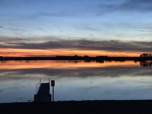 A serene lake at sunset, with a dock extending into calm waters reflecting vibrant orange and blue hues.