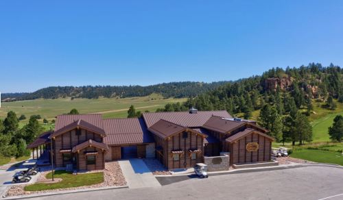 A large wooden lodge with a metal roof, surrounded by green hills and trees under a clear blue sky.
