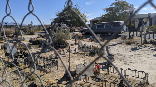 A cemetery scene viewed through a chain-link fence, featuring gravestones, flowers, and an old car in the background.