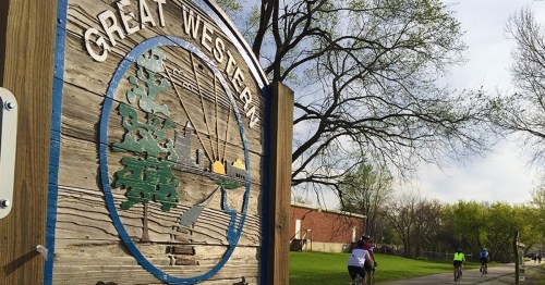 Sign for Great Western Trail with cyclists riding nearby, surrounded by trees and greenery.