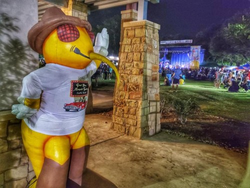 A large, colorful bird statue wearing a hat and shirt stands near a crowd enjoying a nighttime outdoor event.