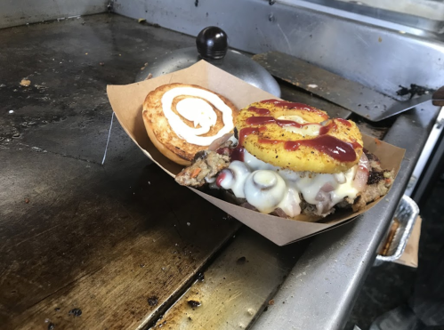 A burger with grilled pineapple, cheese, and condiments on a tray, next to a bun on a griddle.