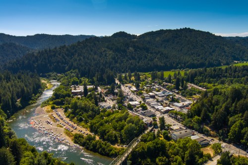 Aerial view of a small town surrounded by lush green hills and a river, with sandy areas and parked cars along the shore.