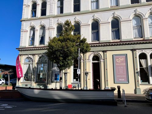 A historic building with large windows, featuring a boat-shaped planter and a "Now Open" banner outside.