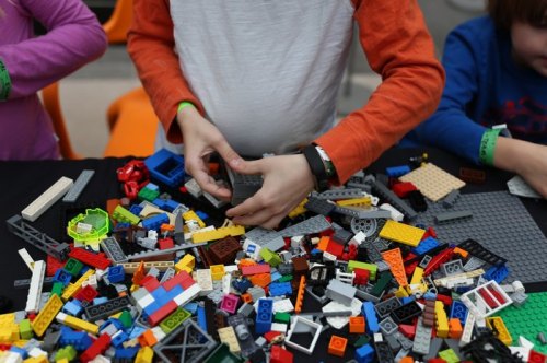 A child’s hands assembling LEGO bricks from a colorful pile on a table, with other children visible in the background.