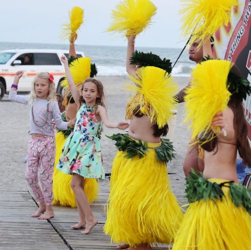 Children in colorful outfits dance on a beach, waving yellow feathered props against a seaside backdrop.