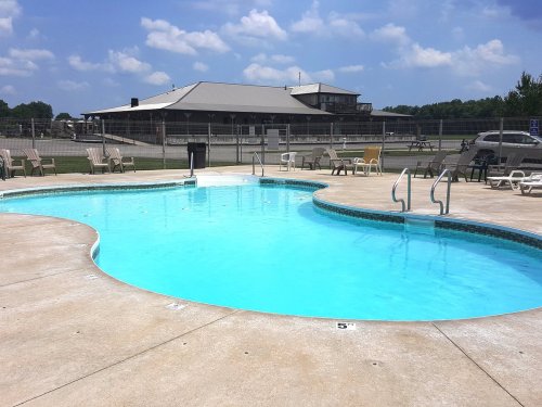A clear blue swimming pool with lounge chairs, surrounded by a grassy area and a building in the background under a blue sky.