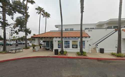 A building with a tiled roof, palm trees, and a parking lot, featuring storefront windows and outdoor seating.