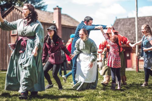 A lively outdoor scene with people in historical costumes dancing and holding hands on a sunny day.