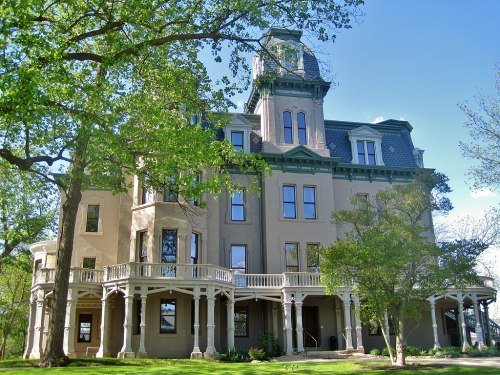 A large historic mansion with a tower, surrounded by trees and greenery under a clear blue sky.