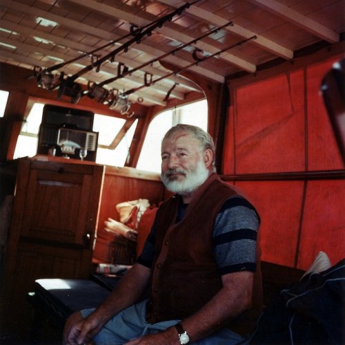 A man with a beard sits inside a boat, surrounded by fishing gear and warm, red-toned interior.