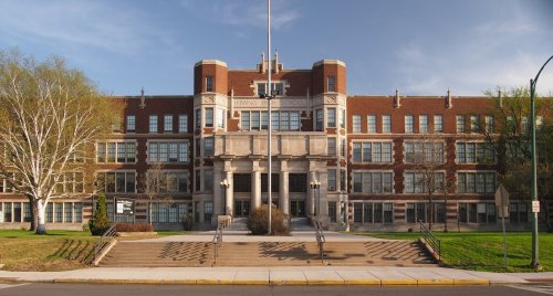 Historic brick school building with large entrance steps and trees, under a clear blue sky.