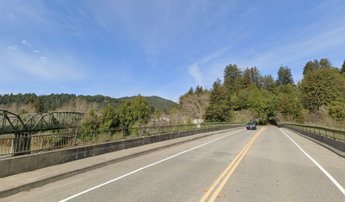 A scenic view of a bridge over a river, surrounded by trees and mountains under a clear blue sky.