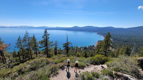 Two hikers walk along a trail overlooking a blue lake surrounded by mountains and trees on a sunny day.