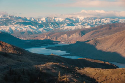A scenic view of snow-capped mountains and a winding river surrounded by rolling hills under a clear sky.