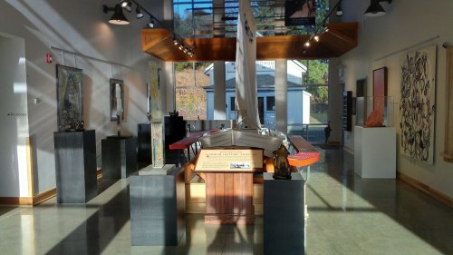 Interior view of an art gallery featuring sculptures, paintings, and a model sailboat under natural light.