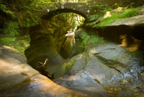 A serene landscape featuring a stone bridge over a narrow, winding creek surrounded by lush greenery and rocky terrain.