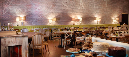 Cozy restaurant interior with wooden tables, green seating, and a display of cakes and desserts against a brick wall.