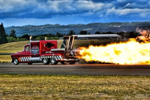 A red truck with a jet engine accelerates on a runway, emitting a large flame against a cloudy sky.