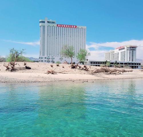 A clear blue lake in the foreground with a sandy shore and a large hotel building in the background under a bright sky.