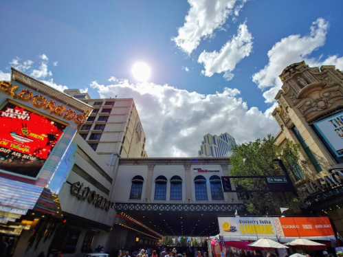 Bright sun shines over a bustling street with colorful signs and people, framed by buildings and a cloudy sky.