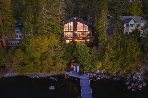 Aerial view of a lakeside cabin surrounded by trees, with a wooden dock leading to the water. Warm lights glow from the windows.
