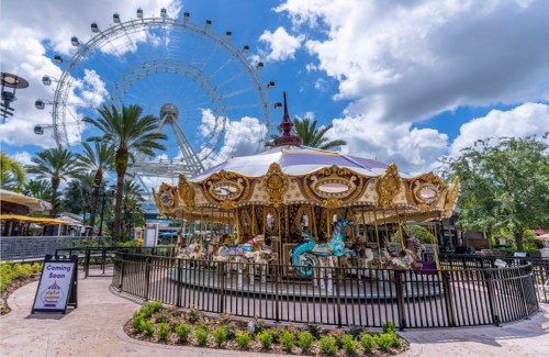 A colorful carousel with ornate horses in front of a large Ferris wheel under a blue sky with fluffy clouds.