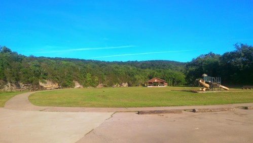 A sunny park with a playground, pavilion, and green grass, surrounded by trees and rocky cliffs in the background.