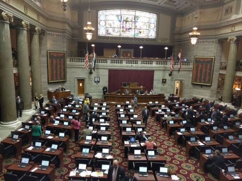 A view of a legislative chamber with desks, computers, and officials engaged in discussion. Stained glass windows above.