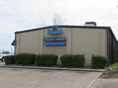 Exterior of a restaurant named "Blue Canoe" with the slogan "good mood food," surrounded by shrubs and a cloudy sky.