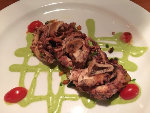 Grilled octopus served on a plate with green sauce, cherry tomatoes, and diced vegetables.