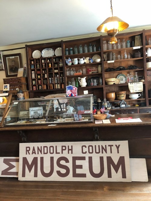Interior of a museum featuring a wooden display counter, shelves with artifacts, and a large sign reading "Randolph County Museum."