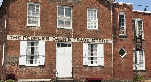 Historic brick building with "The People's Cash & Trade Store" sign, featuring white shutters and a museum sign nearby.