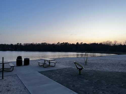 A serene lakeside view at sunset, featuring picnic tables and exercise equipment on a sandy shore.