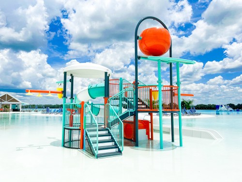 Colorful water play structure with slides and a large orange feature, set against a bright blue sky and fluffy clouds.