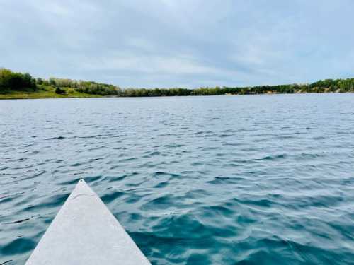 A calm lake viewed from the tip of a kayak, surrounded by green trees under a cloudy sky.