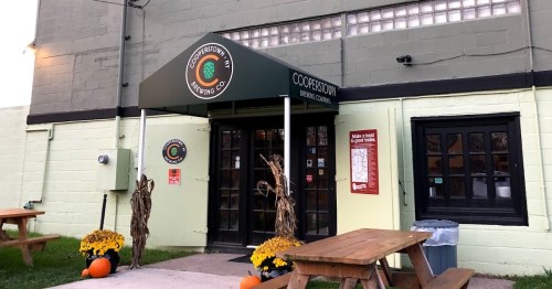 Entrance of Cooperstown Brewing Company with a green awning, picnic tables, and autumn decorations.