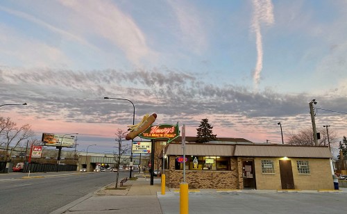 A fast-food restaurant at dusk, with a colorful sky and clouds, featuring a neon sign and nearby billboards.