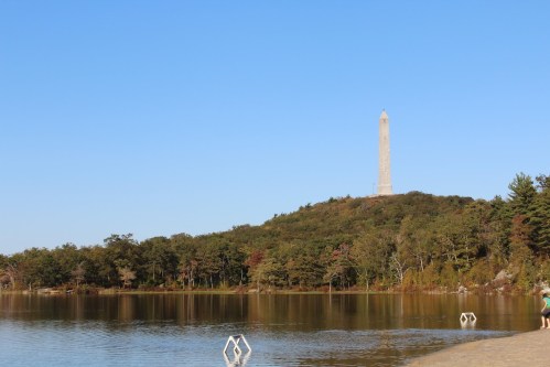 A tall monument on a hill overlooking a calm lake, surrounded by trees under a clear blue sky.