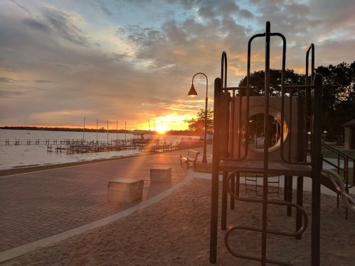 A playground structure silhouetted against a sunset over a lake, with benches and docks in the background.
