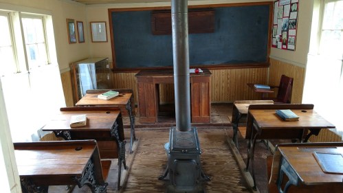 A vintage classroom with wooden desks, a central stove, and a chalkboard, evoking a historical educational setting.