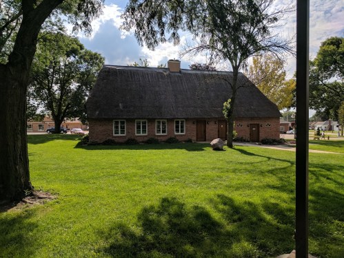 A thatched-roof house surrounded by green grass and trees under a partly cloudy sky.