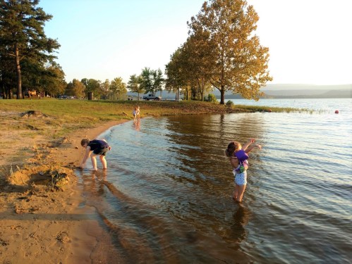 Children playing by the water's edge at sunset, with trees and a calm lake in the background.