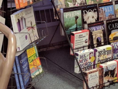 A display rack filled with various books, including travel guides and puzzles, in a cozy bookstore setting.