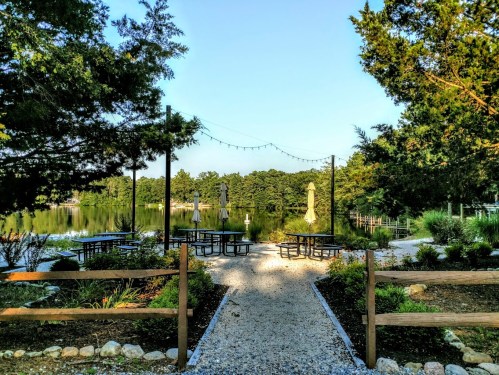 A serene lakeside area with picnic tables, string lights, and lush greenery, framed by wooden fencing.