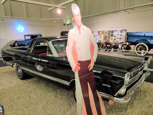 A vintage black car with a life-sized cutout of a man in a white shirt, displayed in a museum setting.