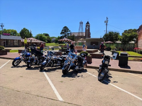 A row of parked motorcycles in a sunny town square with people and a clock tower in the background.