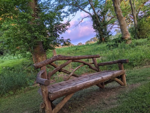 A rustic wooden bench sits in a lush green area, surrounded by trees and a colorful sky at dusk.