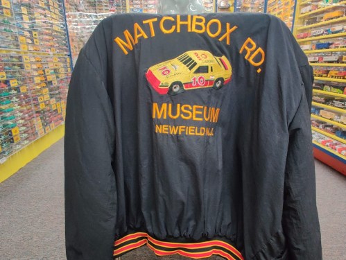 Black jacket featuring "Matchbox Rd. Museum" and an embroidered car design on the back, displayed in a colorful store.