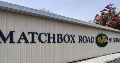 Sign for Matchbox Road Museum, featuring colorful graphics and surrounded by greenery.